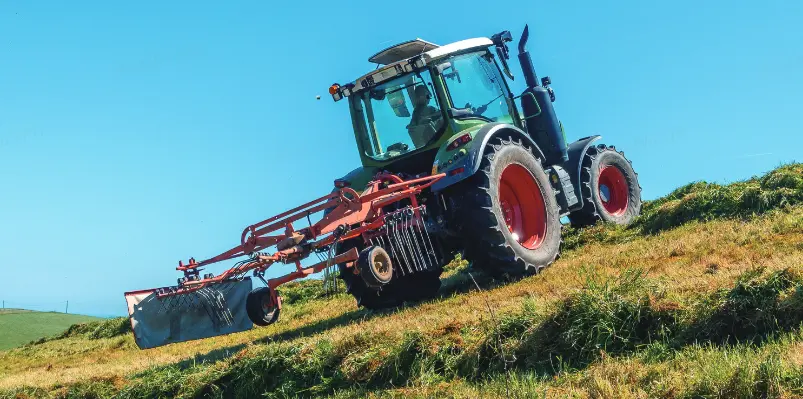 A vibrant green modern tractor with red wheel hubs climbs a grassy slope, pulling a large red hay rake. A driver is visible in the cab. The scene, set against a backdrop of bright blue sky and distant green fields, captures the power and efficiency of agricultural machinery working on hilly terrain during haymaking season.